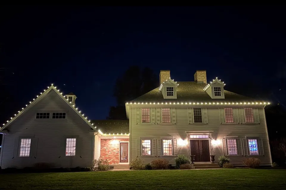 A white colonial with an attached wing, warm white lights along both rooflines and around the dormer windows.