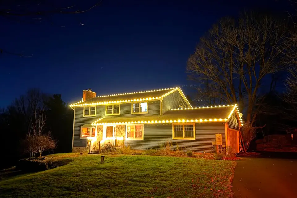 Warm white roofline lights on a two-story colonial against a deep blue dusk sky.