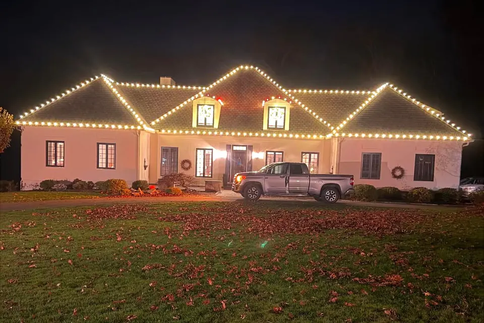 A wide single-story home with warm white lights following every gable peak, seen across a deep front lawn covered in fallen leaves.